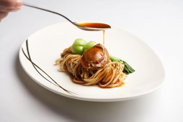 person drizzling sauce on abalone and noodle dish