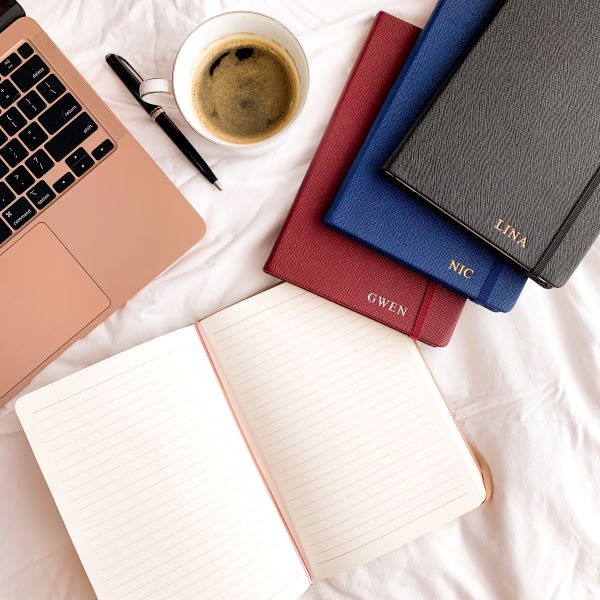 flat lay of leather bound notebooks in black, red, blue, laptop and coffee on white bedsheets
