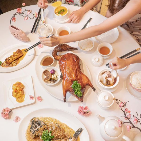 round table spread including dishes like roasted duck and steamed fish with people reaching for them